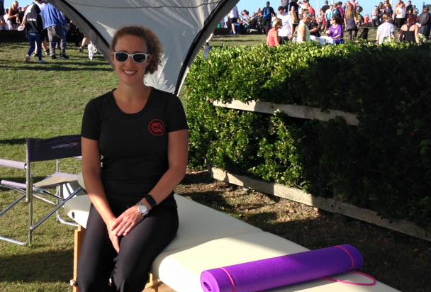 Claire sitting on a portable treatment couch under a white gazebo at an outdoor event, wearing a black polo shirt with her logo